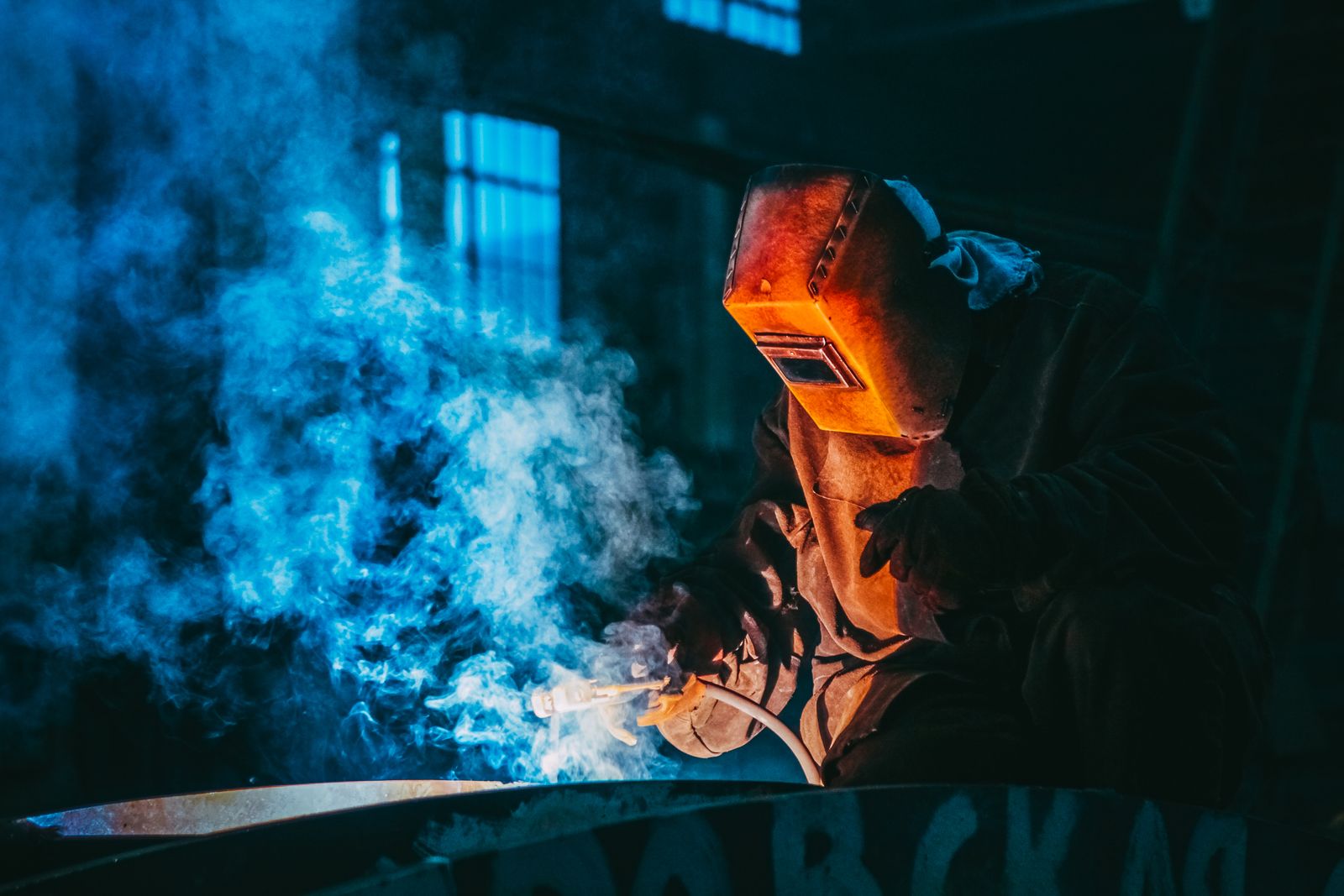 A welder working in a workshop surrounded by blue smoke and sparks