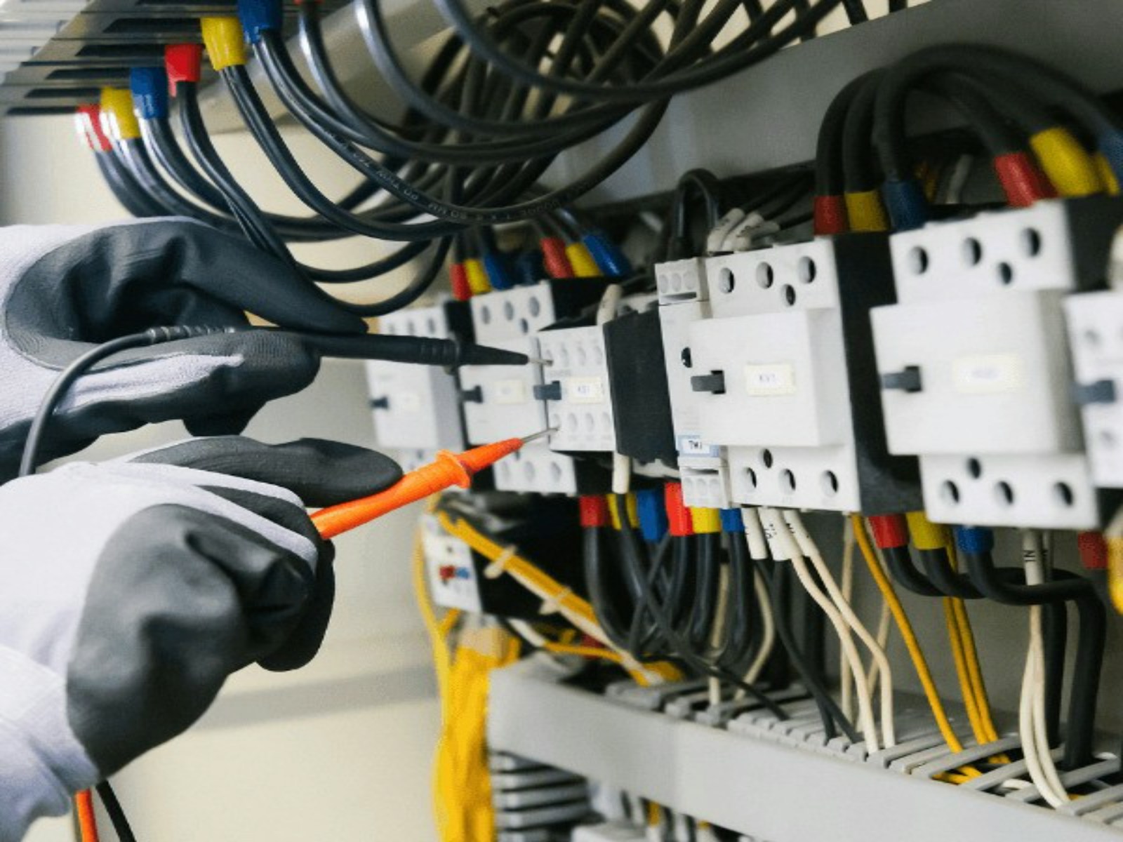 An electrician in insulated gloves testing a control panel with a multimeter probe