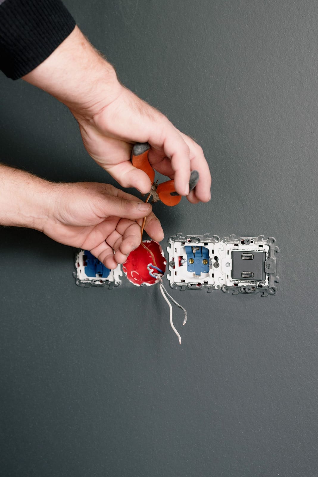 An electrician's hands using pliers to install electrical sockets against a gray wall