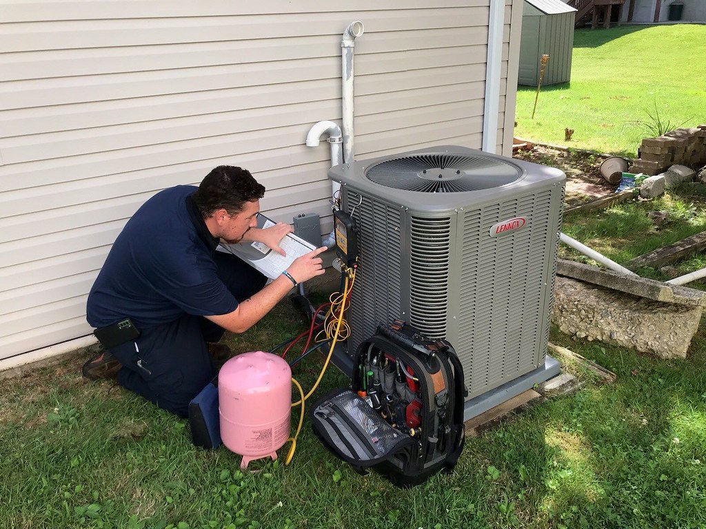 An HVAC technician kneeling beside an outdoor condenser unit with a recovery tank and gauges