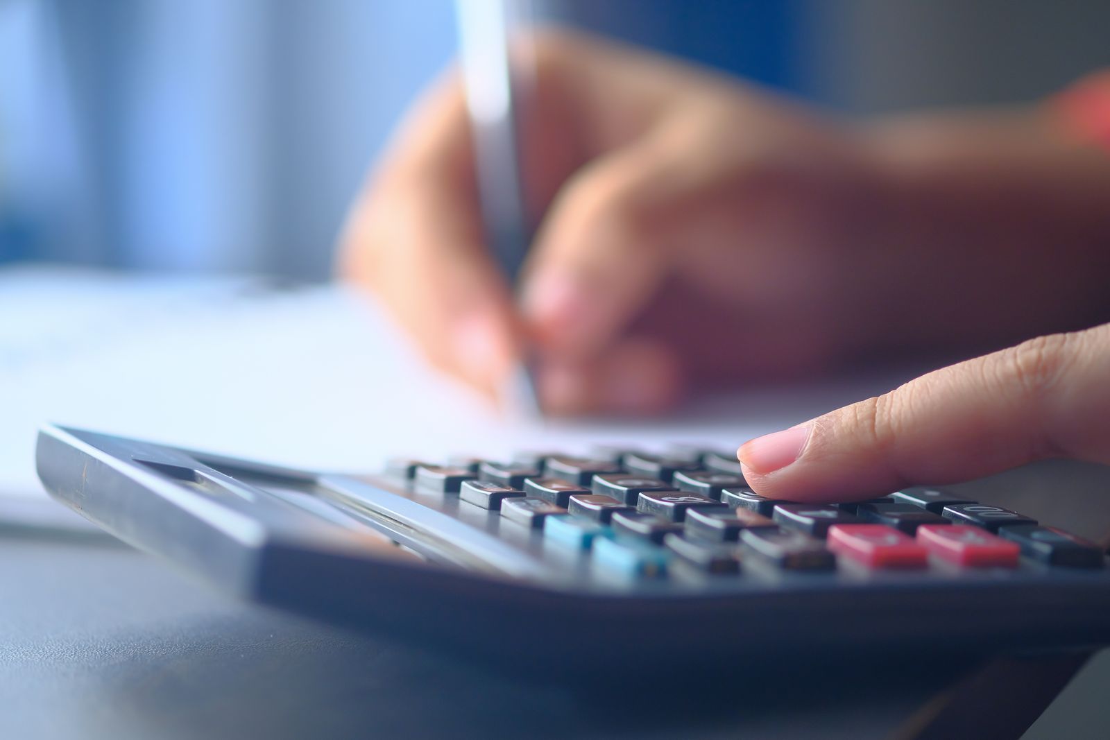 A finger resting on a calculator while the other hand writes a figure on paper.