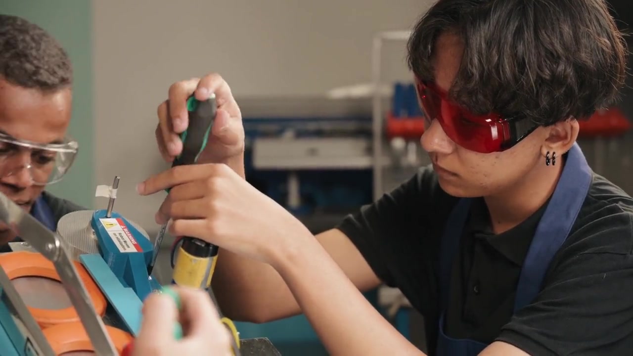 Two trainees in safety glasses working with hand tools at a technical workbench.